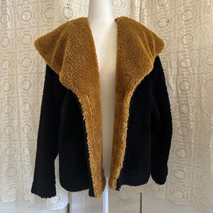 Peut inclure: Veste teddy bear noire et beige clair sur un mannequin. La veste présente un grand col et revers beiges clairs contrastant avec le corps noir. Fabriquée dans une matière douce et duveteuse, idéale pour l'hiver.