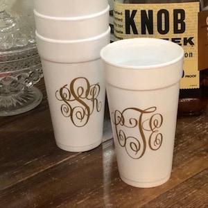 May include: A set of six white plastic cups with gold monogram designs. The cups are stacked on top of each other. The cups are on a wooden surface.