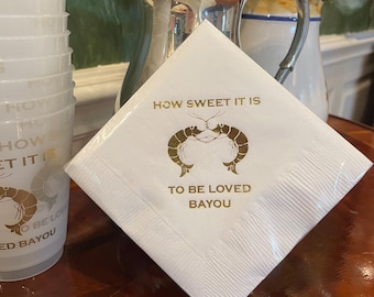 May include: White paper napkin with gold foil lettering and a design of two shrimp. The text reads "How sweet it is to be loved Bayou".