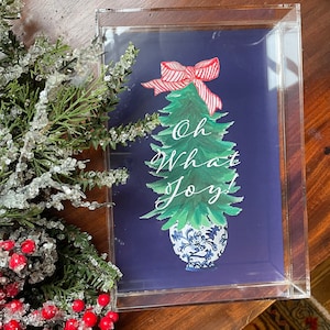 May include: A Christmas-themed print in a clear acrylic frame. The print features a green Christmas tree with a red and white striped bow, a blue and white vase, and the words "Oh What Joy!" on a dark blue background.
