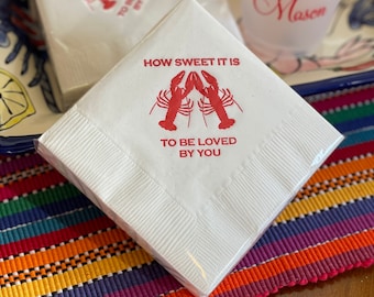 Puede incluir: Servilletas de papel blancas con un diseño de langosta roja y el texto "HOW SWEET IT IS TO BE LOVED BY YOU." También se ve un vaso de plástico transparente con los nombres "Catherine and Mason". Las servilletas están empaquetadas.