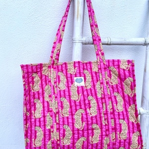 May include: A quilted tote bag in a vibrant pink hue, adorned with a repeating pattern of cheetahs and vertical stripes. The bag features long handles and a small label with text. The bag is hanging on a white ladder.