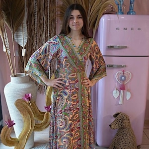 Op de afbeelding: Een vrouw draagt een lange, vloeiende wikkeljurk met een paisley-print in tinten roze, groen, blauw en rood. De jurk heeft een V-hals en lange mouwen. Ze staat voor een roze koelkast met het woord "SMEG" erop.