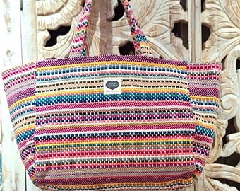 May include: A large, colorful striped tote bag with a woven texture. The bag has a brown leather tag with the text "LILA ZUMBEKO" and a heart symbol.