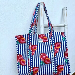 May include: A quilted tote bag with a blue and white striped pattern. The bag is decorated with a repeating pattern of red pomegranates. The handles are striped with red, blue, and white. The bag is hanging on a white ladder.