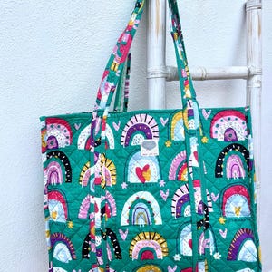 May include: A quilted tote bag with a vibrant rainbow pattern on a teal background. The bag features a repeating design of colorful rainbows, hearts, and stars. The handles and trim are a coordinating multi-colored pattern. The bag is displayed against a white background.
