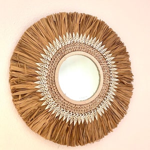 Puede incluir: Un espejo redondo con un marco tejido natural y un borde decorativo de conchas blancas. El espejo tiene una estética rústica y costera.