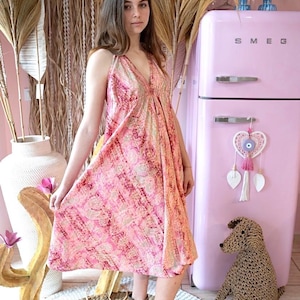 Może przedstawiać: Kobieta w różowej sukience z wzorami w kolorze złotym i dekoltem w kształcie litery V. Stoi przed różową lodówką z napisem "SMEG".