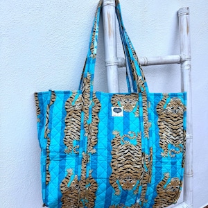 May include: A quilted tote bag with a vibrant blue and white striped pattern. The bag features a repeating design of brown and black tiger illustrations. The bag has long blue straps and a small label with text.