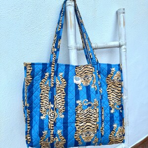 May include: Blue quilted tote bag with vertical stripes and a repeating tiger print in shades of tan and black. The bag has matching blue handles and a small tag with a heart design.