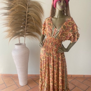 May include: A long, flowing maxi dress with a paisley print in shades of orange, yellow, and green. The dress has a V-neckline, short sleeves, and a fitted waist. The model is wearing a pink head wrap.
