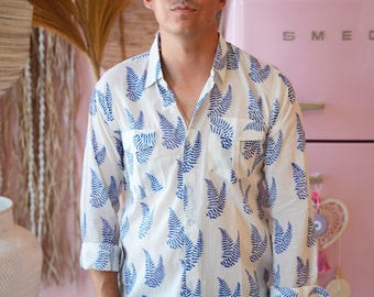 May include: White button-down shirt with a blue fern pattern. The shirt has long sleeves, a chest pocket, and is paired with blue jeans. The background includes a pink refrigerator and decorative items.