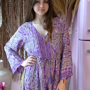 Puede incluir: Un vestido largo de manga larga de color morado con un lazo en la cintura y un diseño estampado en dorado y morado. El vestido tiene cuello en V y una falda fluida.