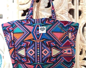 May include: A large tote bag with a colorful geometric pattern in shades of pink, blue, orange, and green. The bag has a black background and a small heart-shaped tag with the text "Lola Tribe" on it.