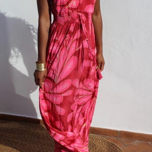 May include: A floor-length sleeveless dress in shades of deep red and bright pink, featuring a repeating abstract petal pattern with gold outlines. The garment has a V-neckline, thin spaghetti straps, and a gathered, elasticised waistline.