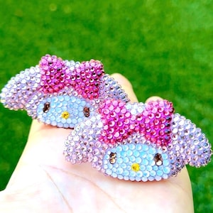 May include: Two hair clips shaped like the character My Melody, fully covered in sparkling rhinestones. The design features a light purple face, dark brown eyes, a yellow nose, and a bright pink bow on top, all crafted with small crystals.