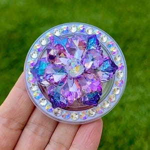 May include: A round, bedazzled phone grip with a floral design. The center features a cluster of purple and blue crystals, surrounded by a ring of smaller, clear crystals. The grip is held against a blurred green background.