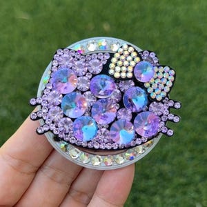 May include: A bedazzled phone grip featuring a Hello Kitty design. The grip is adorned with iridescent rhinestones in shades of purple, blue, and clear, set against a black outline. The grip is round with a silver rim.