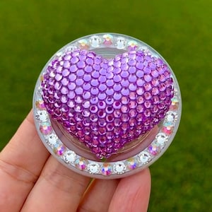 May include: A bedazzled phone grip featuring a purple heart design. The heart is covered in small, round, purple rhinestones. The grip is surrounded by a ring of clear, iridescent rhinestones, set in a clear plastic base. The background is a blurred green.
