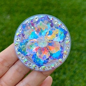 May include: A clear, circular phone grip with a sparkling, multi-colored flower design. The flower is made of iridescent crystals, surrounded by a ring of smaller, clear crystals. The grip is held in a hand against a blurred green background.