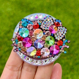 May include: A bedazzled phone grip featuring a cat face design. The grip is adorned with a variety of colorful rhinestones, including blue, green, pink, and red. The grip has a silver border and is set against a green background.