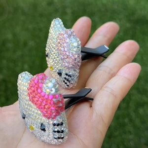 May include: Two Hello Kitty car vent clips, encrusted with iridescent rhinestones. The clips feature a silver base, black accents, and a pink heart. The rhinestones reflect a rainbow of colors. The clips are held in a hand against a blurred green background.