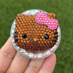 May include: A bedazzled phone grip featuring a cartoon cat face with a pink bow. The cat's face is made of orange rhinestones, with black eyes and a gold nose. The grip is surrounded by clear and iridescent rhinestones.