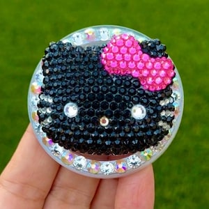 May include: A bedazzled phone grip featuring a black cat face with a pink bow. The cat's face is made of black rhinestones, with clear rhinestones around the edge. The bow is made of pink rhinestones.