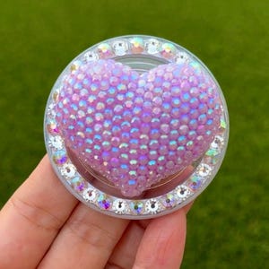 May include: A round, clear phone grip with a heart-shaped design. The heart is made of small, iridescent purple beads. The grip is encircled by clear, sparkling rhinestones, creating a glamorous accessory.