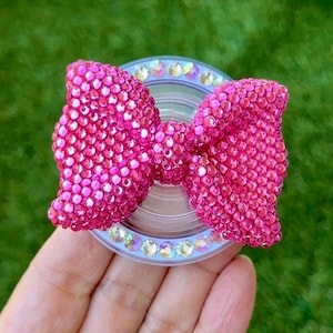 May include: A pink bow-shaped phone grip covered in small, sparkling rhinestones. The grip is attached to a clear, circular base with a ring of iridescent gems. The background is a blurred green.