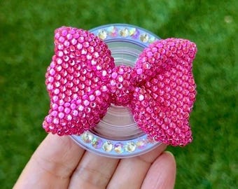 May include: A pink bow-shaped phone grip covered in small, sparkling rhinestones. The grip is attached to a clear, circular base with a ring of iridescent gems. The background is a blurred green.