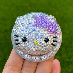 May include: A bedazzled phone grip featuring a Hello Kitty design. The grip is round and covered in iridescent rhinestones, with a purple bow and black eyes. The design is set against a green background.