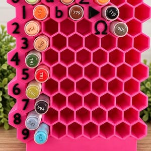 Könnte beinhalten: Ein leuchtend pinker, wabenförmiger Marker-Organizer mit einem Gitter aus sechseckigen Fächern. Die linke Seite ist mit schwarzen Zahlen von 0 bis 9 beschriftet, während die oberste Reihe Buchstaben und Symbole enthält. Marker stecken in den Fächern.