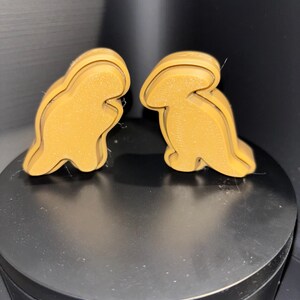 May include: Two brown dinosaur-shaped cookie cutters. The cookie cutters are made of plastic and have a smooth, glossy finish.