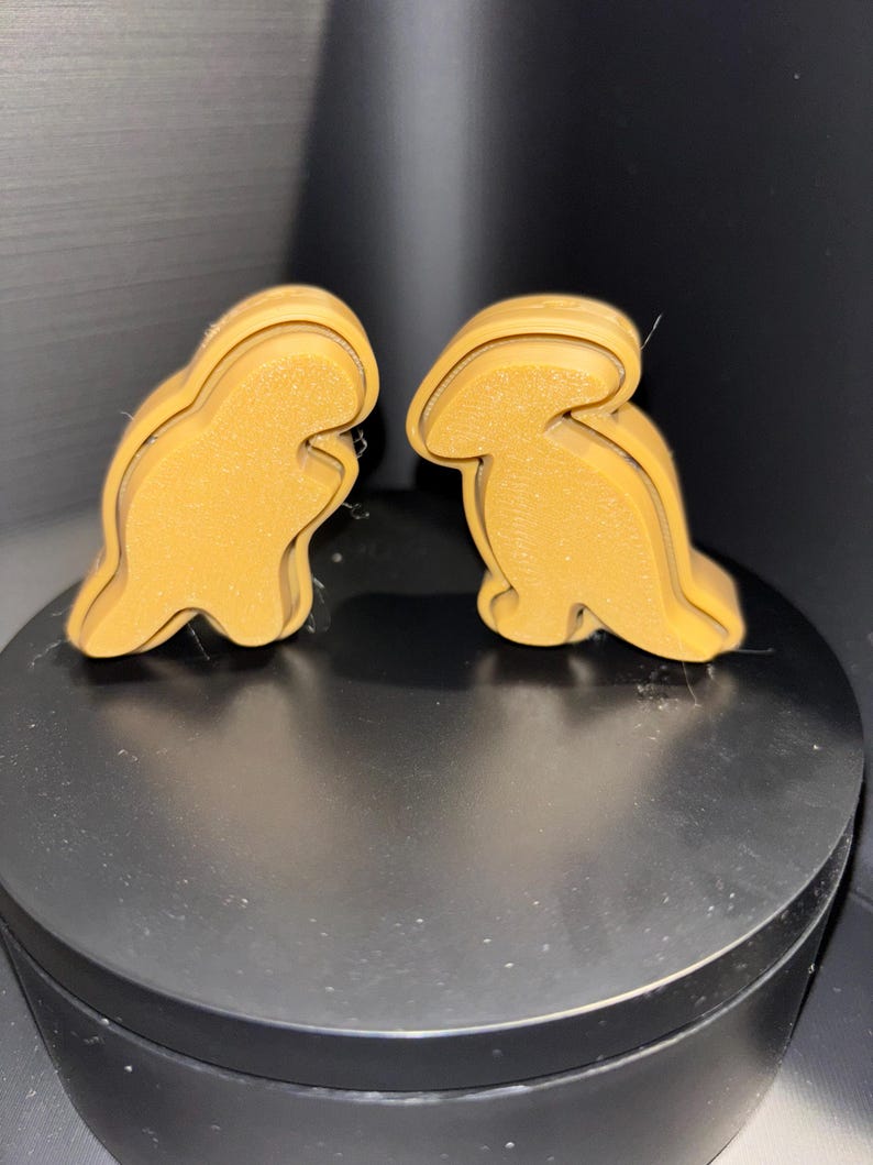 May include: Two brown dinosaur-shaped cookie cutters. The cookie cutters are made of plastic and have a smooth, glossy finish.