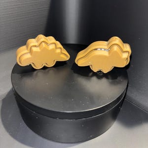 May include: Two brown dinosaur-shaped cookie cutters. The cutters are made of a smooth, shiny plastic.