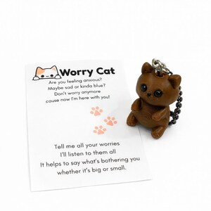 May include: A brown 3D-printed cat figurine keychain with large black eyes and a metal chain, accompanied by a white card featuring a cat illustration, orange paw prints, and the text: Worry Cat, Are you feeling anxious? Maybe sad or kinda blue? Don't worry anymore cause now I'm here with you! Tell me all your worries I'll listen to them all It helps to say what's bothering you whether it's big or small.