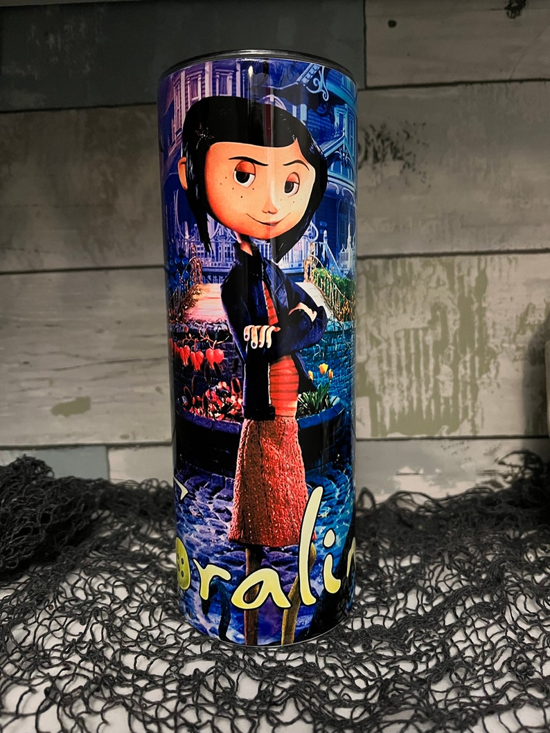 May include: A stainless steel tumbler with a colorful image of a girl with black hair and a red dress. The image is from the movie Coraline. The tumbler has a black lid and a black straw.