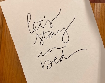May include: A white rectangular piece of paper featuring the dark gray handwritten cursive text let's stay in bed. The lettering is arranged in four lines with a flowing script style, centered on the page against a plain white background.