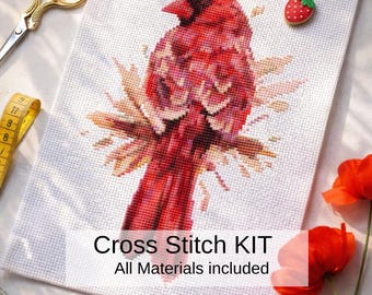 May include: A cross-stitch kit featuring a vibrant red cardinal bird design. The kit includes all materials and is displayed with scissors, a measuring tape, and a strawberry-shaped pin. The text "Cross Stitch KIT" and "All Materials included" is visible.