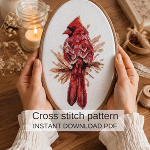 May include: A cross-stitch pattern featuring a red cardinal bird design, displayed in an oval wooden embroidery hoop. The text "Cross stitch pattern INSTANT DOWNLOAD PDF" is visible at the bottom of the image. The bird is surrounded by brown leaves.