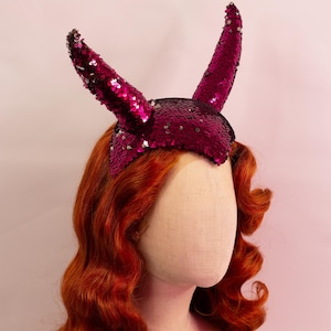 Könnte beinhalten: Ein Schaufensterpuppenkopf mit langem, welligem, rotbraunem Haar und einem paillettenbesetzten Stirnband mit zwei aufrechten Hörnern. Die Hörner und das Stirnband sind tiefmagenta. Der Hintergrund ist ein zartes Rosa.