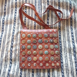 May include: A brown leather crossbody bag with a square shape. The bag is decorated with numerous metal discs and studs. It has a long, adjustable brown leather strap. The bag is set against a striped fabric background.