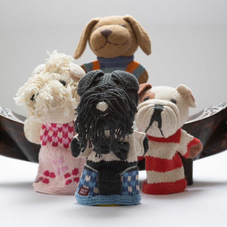 Pu&ograve; includere: Quattro marionette per dita a forma di cane in maglia. Le marionette sono in fila, con un cane marrone dietro, un cane bianco davanti, un cane nero al centro e un cane bianco con strisce rosse sulla manica davanti.