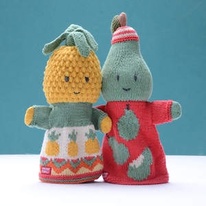 May include: Two knitted fruit-shaped dolls: a yellow pineapple and a green pear. The pineapple doll has a green top and a white skirt with pineapple designs. The pear doll wears a red outfit with pear patterns. Both have smiling faces.