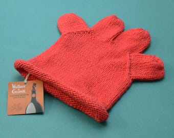 May include: A bright red knitted hat shaped like a hand with four fingers and a thumb, featuring a rolled brim. A square cardboard tag is attached by a string, displaying the text Wallace & Gromit Feathers McGraw Child's Hat and an illustration.