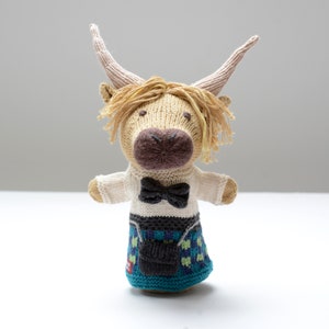 May include: A knitted plush toy of a Highland cow wearing a white shirt with a black bow tie and a blue and green kilt.
