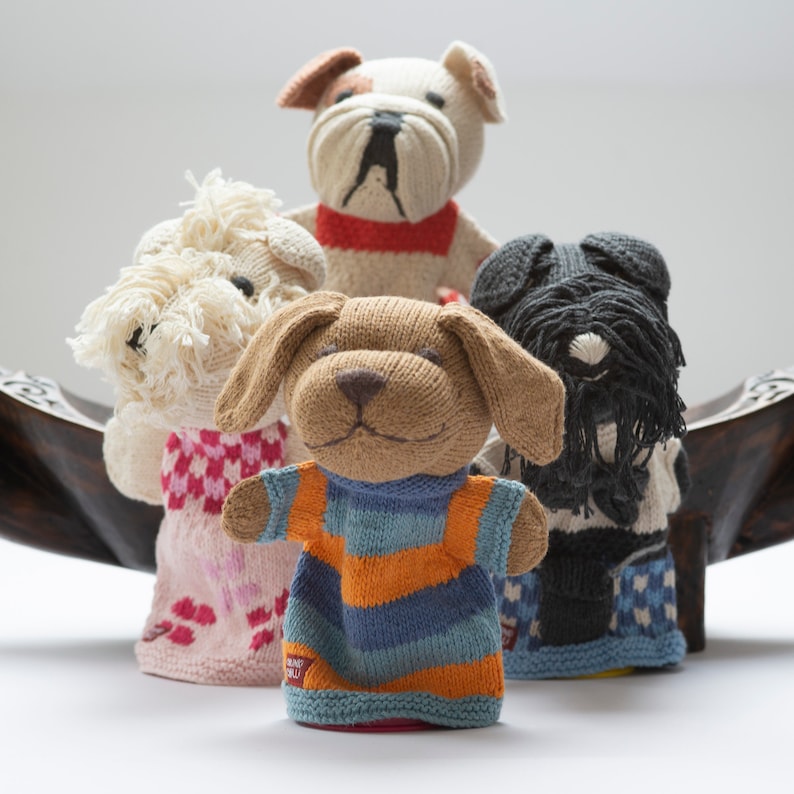 Pu&ograve; includere: Quattro pupazzi per dita di lana a forma di cane. I pupazzi indossano vestiti di diversi colori. I pupazzi sono seduti su una superficie di legno.