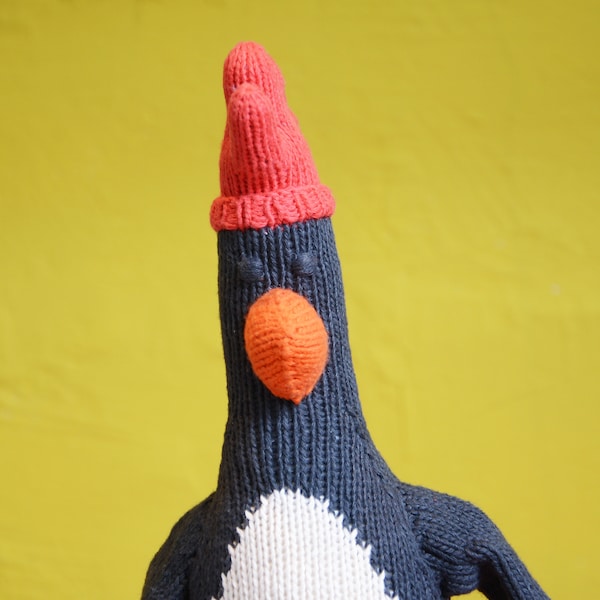 May include: A knitted penguin toy wearing a red knitted hat. The penguin has a black body, white belly, and an orange beak.