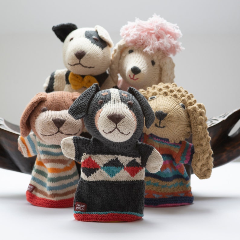 Pu&ograve; includere: Cinque pupazzi per dita di lana a forma di cane. I pupazzi sono tutti di colori diversi e hanno motivi diversi. I pupazzi sono seduti su una superficie di legno.